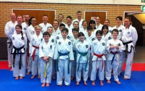 A diverse group of people in martial arts uniforms stand together on a blue mat in the dojo, having spent all day training. The primarily white uniforms display a pattern of colorful belts. A brick wall and two wooden doors provide the backdrop as everyone smiles warmly. - HSTS Taekwon-Do