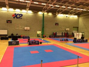 A gymnasium with two martial arts competition mats in blue and red hosts the British Championship. Chairs and tables with event branding surround the mats. Two basketball hoops are mounted on the walls, and a large logo is visible above, commemorating UKITF achievements. - HSTS Taekwon-Do