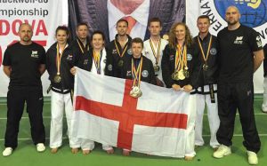 A group of people in martial arts uniforms stand proudly on a green surface, some holding medals. They display an English flag. A large banner and the ITF logo for the 2015 European Taekwondo Championships are in the background. - HSTS Taekwon-Do