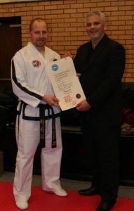 Two men are standing indoors on a red mat. The man on the left, wearing a white martial arts uniform and black belt, is holding a large certificate with an expression of surprise. The man on the right, in a dark suit, is smiling during the presentation of the certificate. - HSTS Taekwon-Do