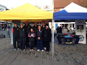 A group of nine people, wearing casual winter clothing, stand smiling under yellow and blue tents at an outdoor market place in Hitchin. A table with Taekwondo materials and a large photo is visible to the right. The setting is a cobblestone plaza, creating a charming backdrop. - HSTS Taekwon-Do