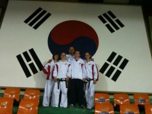 Five people in martial arts uniforms stand in front of a large South Korean flag at the 2010 World Championships. They are wearing white ITF uniforms with black belts, and some have red or blue logos. Orange seats are visible in the foreground. - HSTS Taekwon-Do
