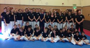 A large group, including adults and children, dressed in martial arts uniforms, pose joyfully in a gym with brick walls and blue mats. The children sit in front while the adults stand behind. Everyone smiles brightly, feeling thankful for Sarah Stevensons guidance. - HSTS Taekwon-Do