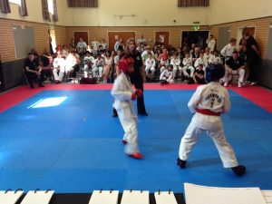 Two young martial artists in sparring gear face each other in a dojo during the HSTS Interclub event, with a referee overseeing the match. Spectators sit around the room, and a judges table with papers is visible at this thrilling Interclub 22-06-14 event. - HSTS Taekwon-Do