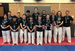 A group of 14 UKITF medalists from 2018, wearing black shirts and white martial arts pants, stand proudly on a red mat. They smile while holding various trophies and medals, with a brick wall and part of a board visible in the background, celebrating their achievement as UK champions. - HSTS Taekwon-Do