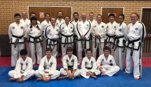 A group of people wearing white martial arts uniforms with black belts stand in rows, posing for a group photo in a gymnasium. Guest Instructor Mr. McCabe, a VIth Degree master, is among them. The background features a brick wall and wooden doors, while some individuals kneel in front. - HSTS Taekwon-Do