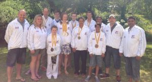 A group of twelve people, from adults to children, stand outdoors in a park-like setting. Theyre wearing white jackets adorned with medals and smiling broadly. It seems theyre celebrating their triumph at the ITF European Championships 2013, surrounded by lush trees and grass. - HSTS Taekwon-Do