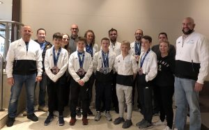 A group of young athletes in white tracksuits, proudly displaying their medals, stand indoors at the 2018 World Championships. Flanked by smiling adults in casual attire, they pose for a photo in what seems to be a lobby or lounge area, capturing their triumph for Argentina. - HSTS Taekwon-Do
