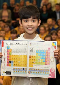 A child grins while holding open a book featuring a colorful periodic table, as if sharing the latest comet news. In the background, a blurred audience is seated, captivated by this young explorers enthusiasm. - HSTS Taekwon-Do
